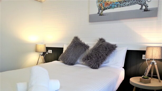 A neatly made bed with white linens, two fluffy pillows, and a folded towel on the bed. There are two bedside tables, each with a lamp. A painting of a leopard is on the wall above the bed. - Kurri Motor Inn (Kurri Kurri)
