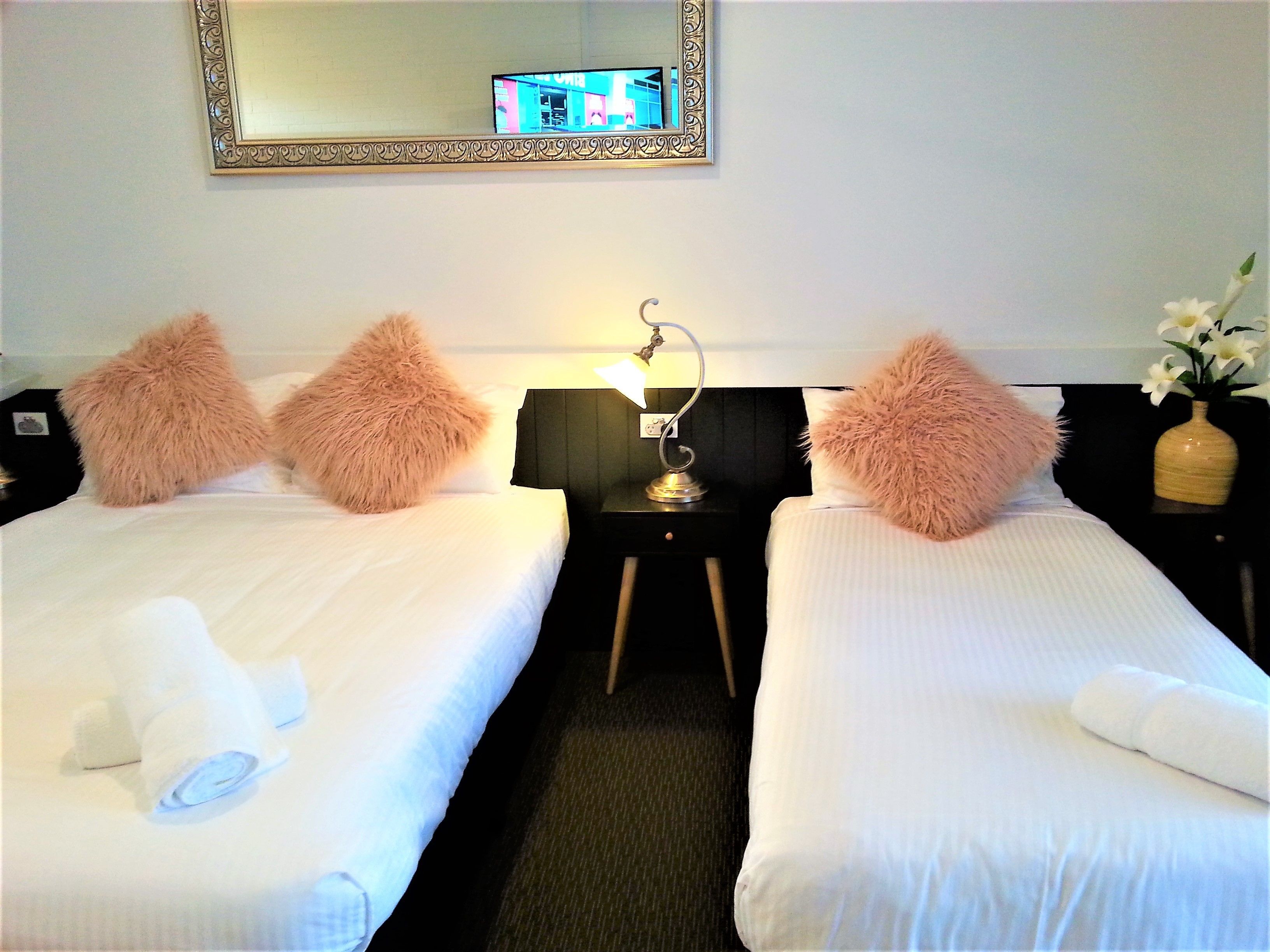 A hotel room with two beds, each with fluffy pillows and neatly folded towels. A small table with a lamp is between the beds.