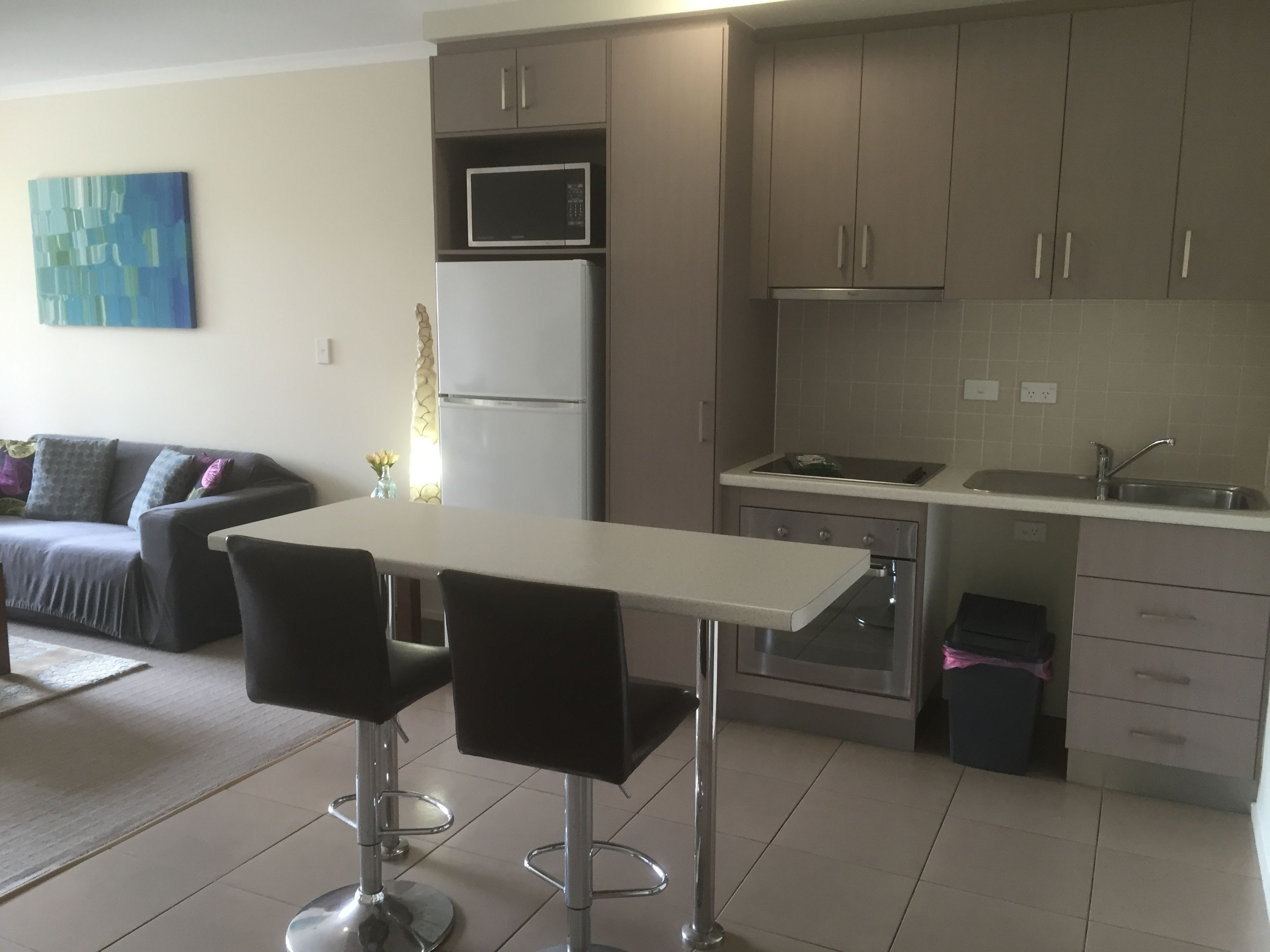 A modern kitchen with a white countertop, stainless steel appliances, and grey cabinets. There is a dining area with a white table and two black chairs.