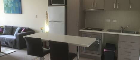 A modern kitchen with a white countertop, stainless steel appliances, and grey cabinets. There is a dining area with a white table and two black chairs.