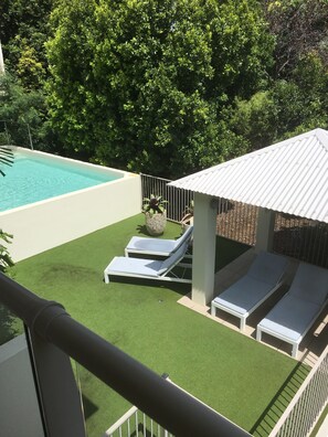 A rooftop pool area with artificial grass, two lounge chairs, and a small structure with a white roof.