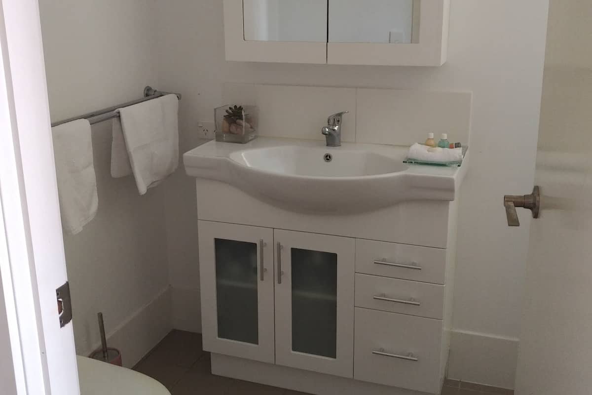 Casa de banho pequena e limpa, com lavatório branco, espelho e armário, sanita e suporte para toalhas.