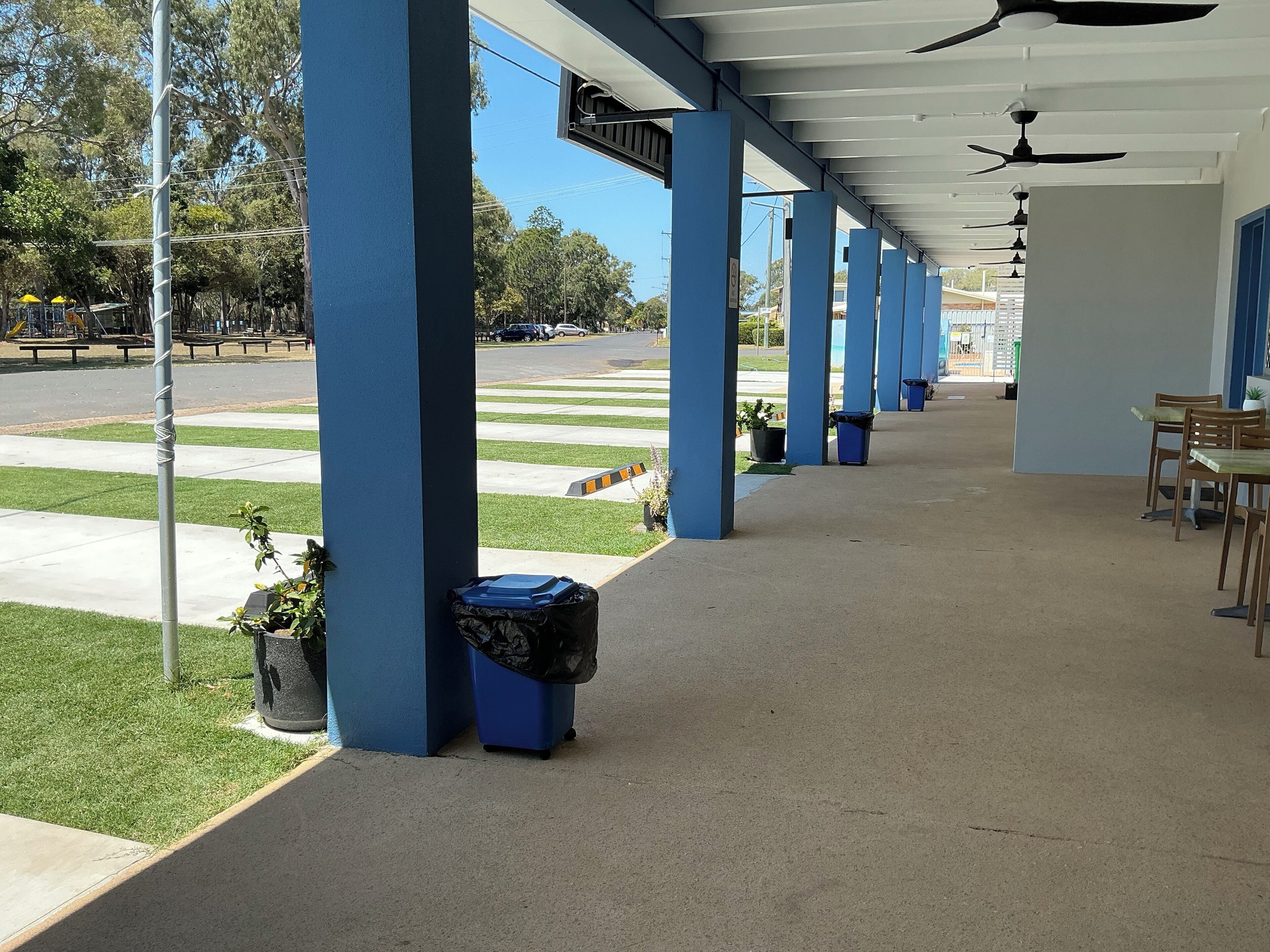 Un espacio al aire libre cubierto con pilares azules, una fila de ventiladores de techo y una zona de estar con mesas y sillas.