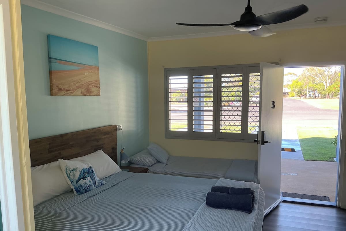 Un dormitorio con cama, mesita de noche, ventilador de techo, ventana con persianas y puerta que da al exterior.
