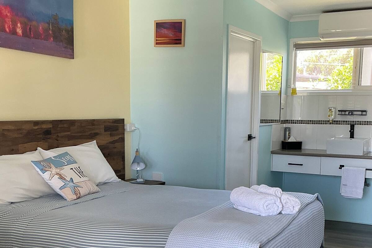 Un dormitorio con una cama grande, una mesita de noche, una lámpara, un cuadro y un baño con lavabo y espejo.