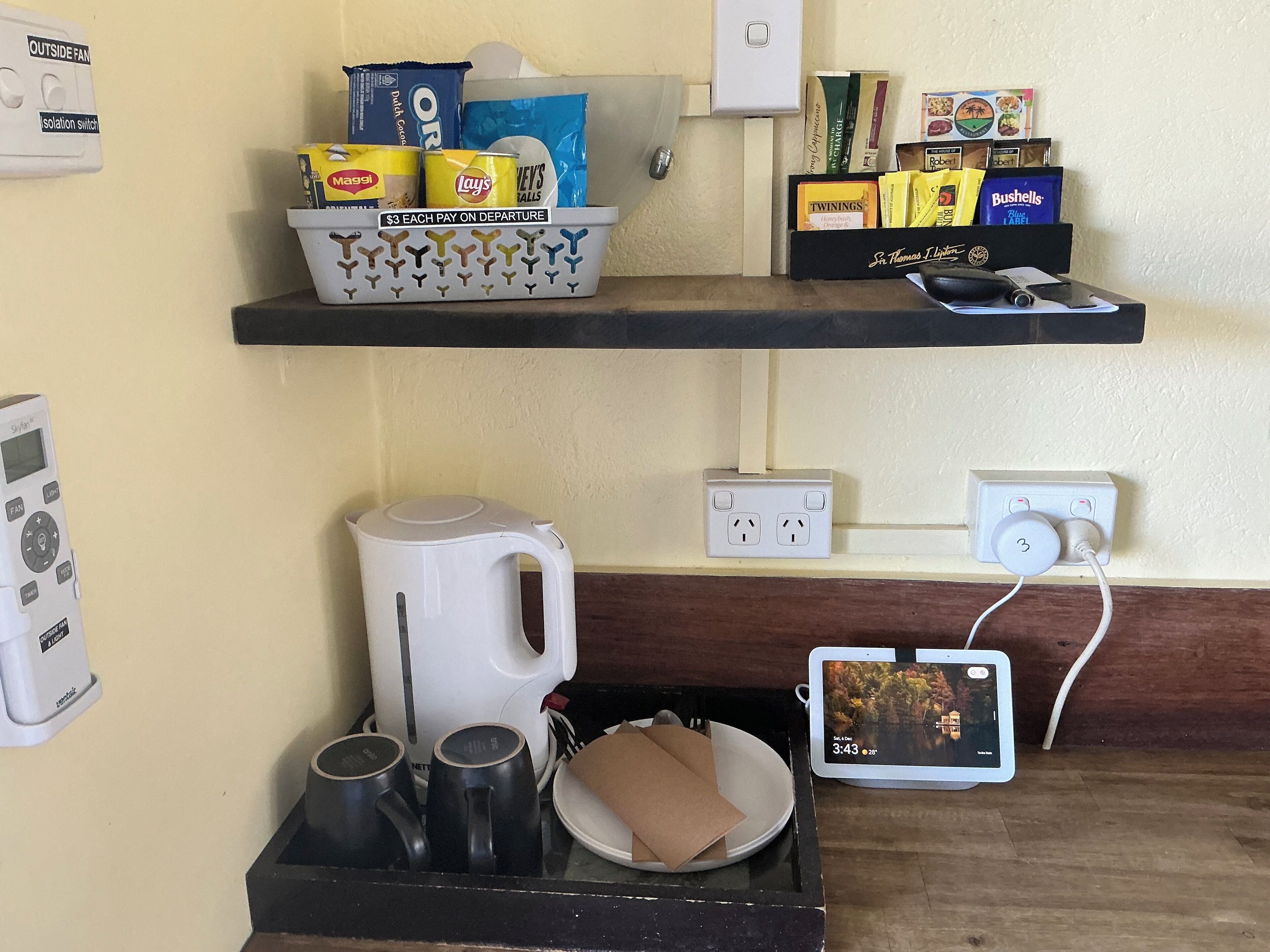 A hotel room with a shelf containing snacks, a kettle, mugs, and a tablet displaying a scenic image.