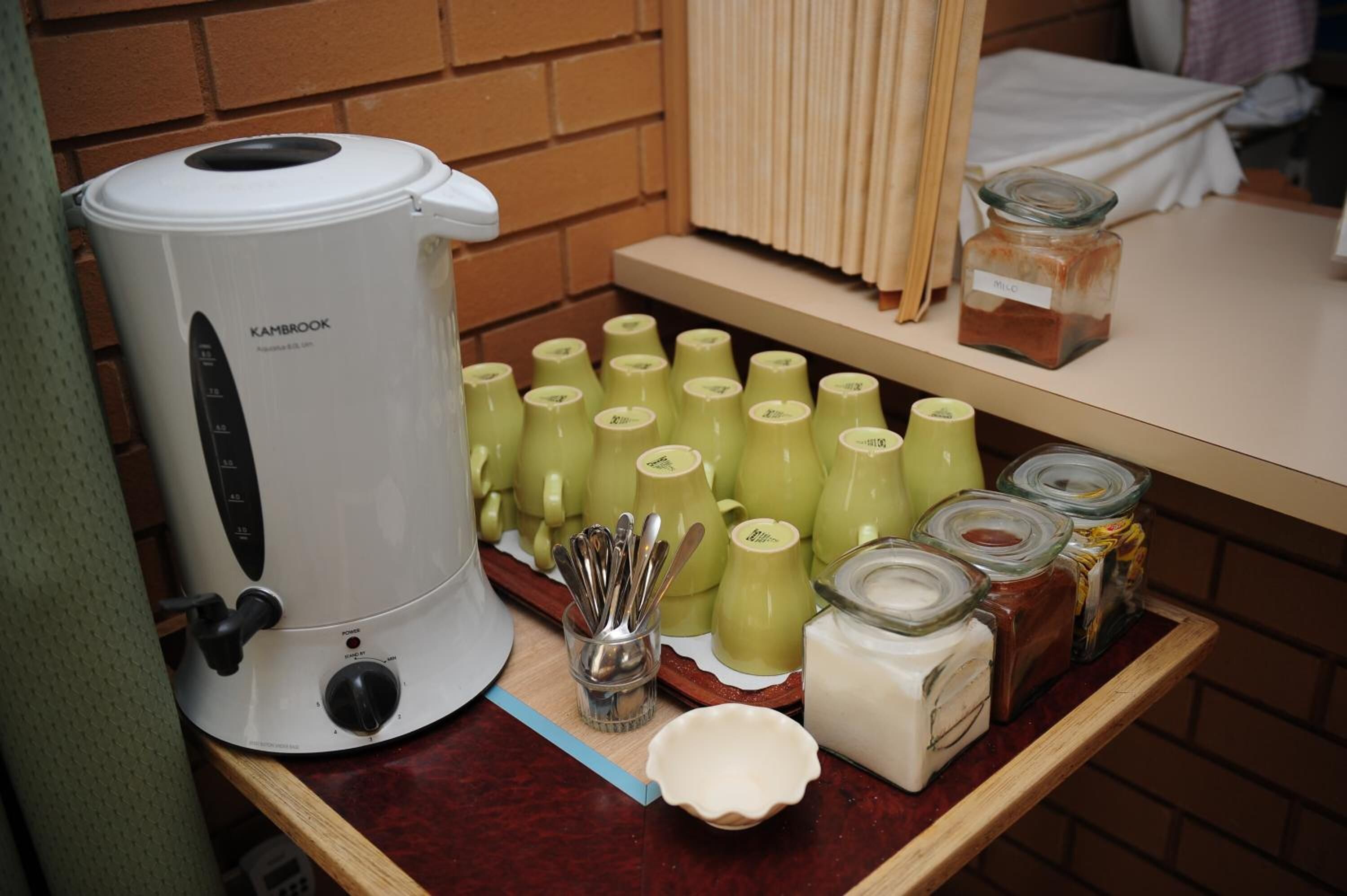Ein Küchenbereich mit einem weißen Wasserkocher, mehreren grünen Tassen und verschiedenen Gläsern mit Gewürzen auf einem Holzregal.