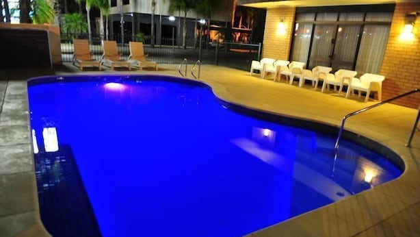 Piscina com tonalidade azul, rodeada por um deck revestido e cadeiras de plástico branco.
