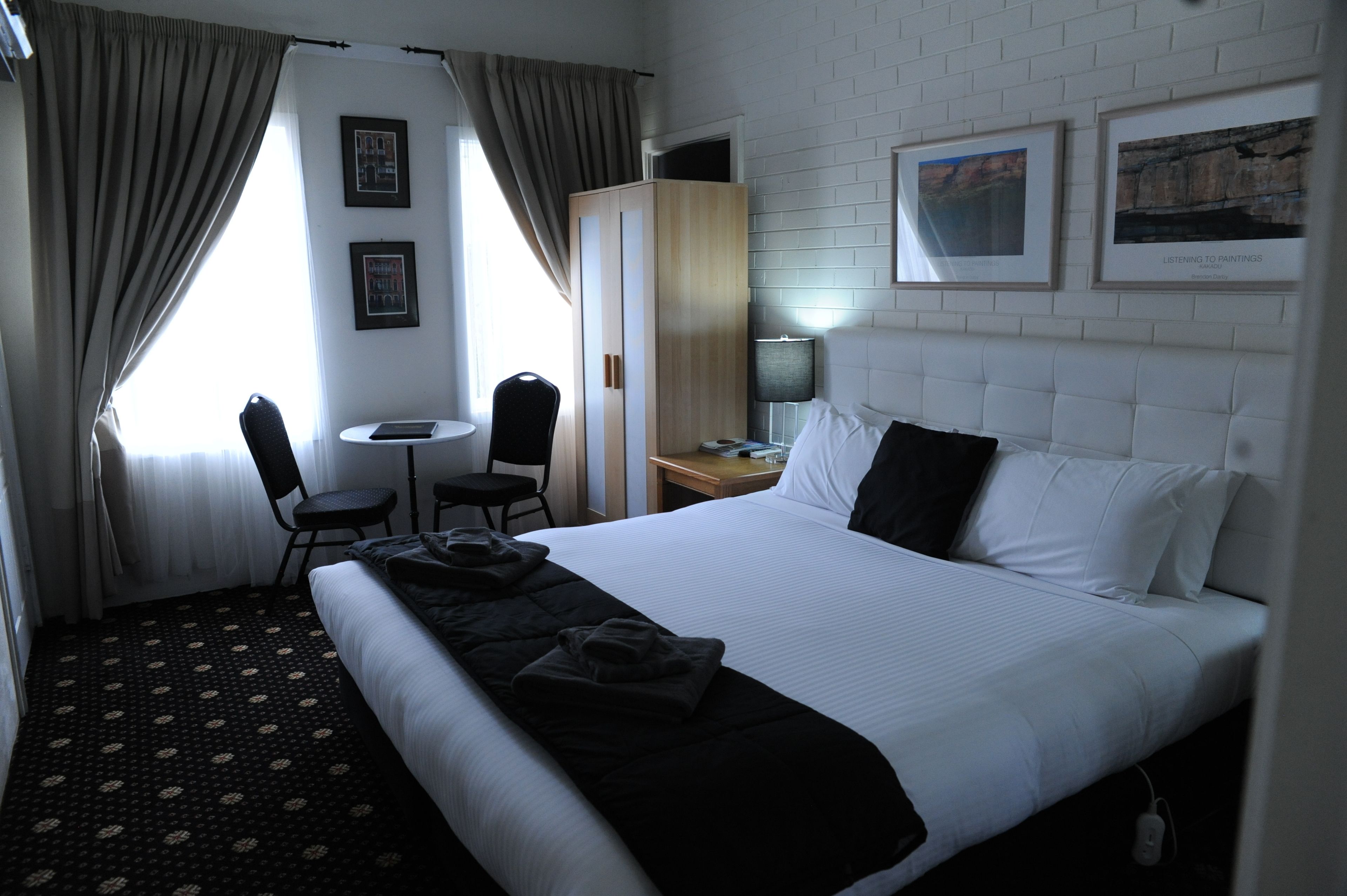Habitación de hotel con una cama grande, un escritorio, una silla y un armario. Hay cuadros enmarcados en la pared y una ventana con cortinas.