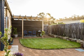 A backyard with a wooden pergola, two blue metal chairs, a barbecue grill, and a fenced area. - Daisy Bank Cottages (Richmond)