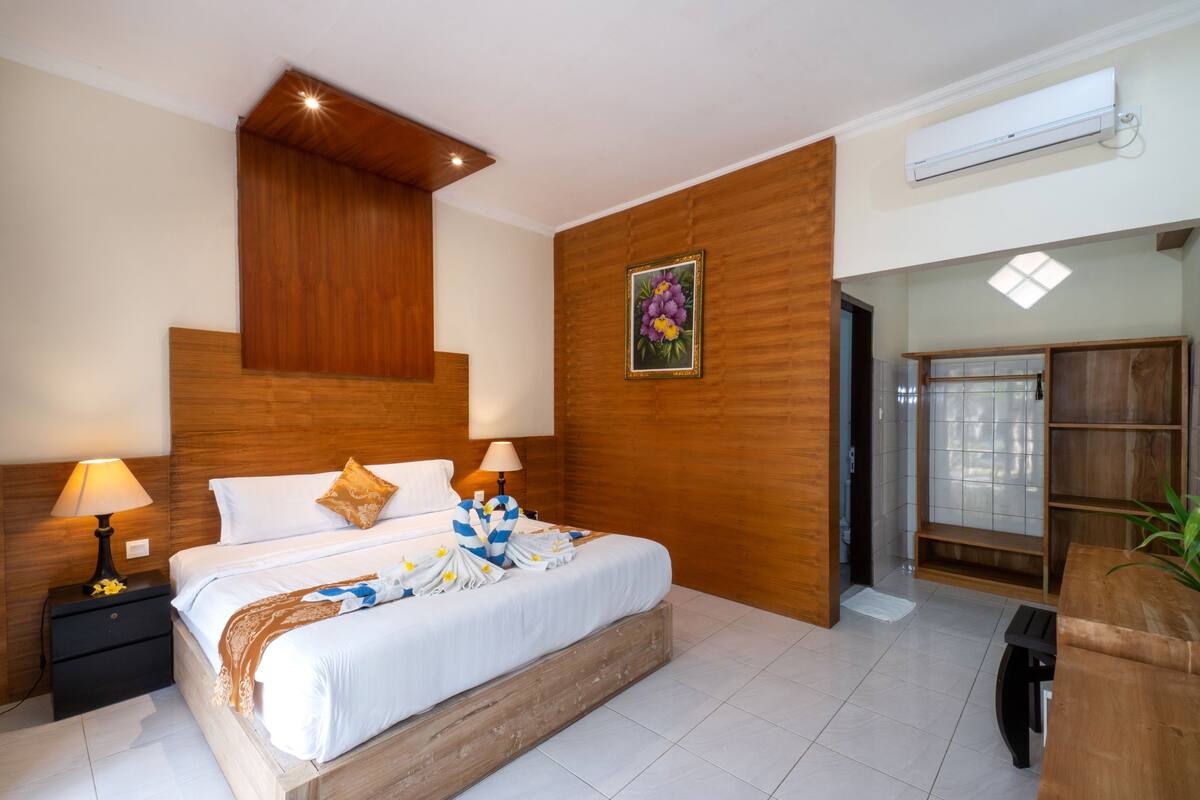 A hotel room with a wooden headboard, a bed with white linens, two bedside lamps, a wooden nightstand, a wooden bench, and a wall-mounted air conditioner.