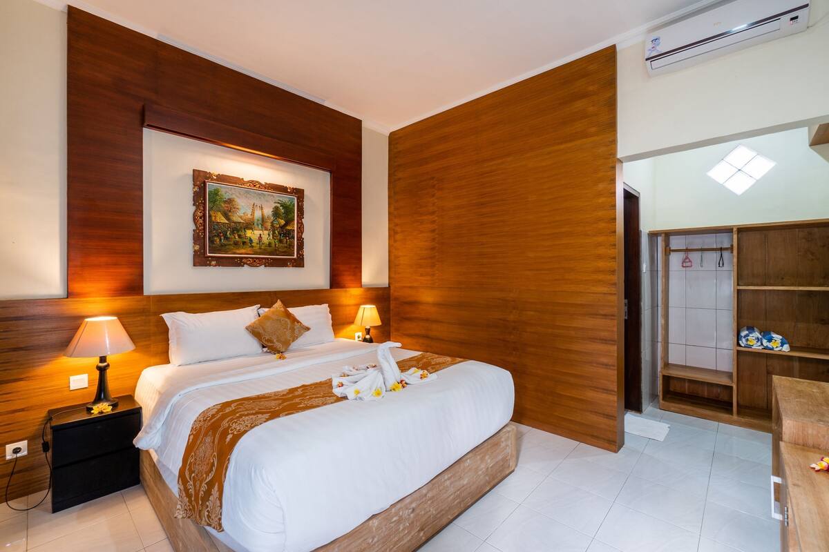 A hotel room with a large bed, wooden headboard, a painting on the wall, a nightstand with a lamp, and a wooden wardrobe.