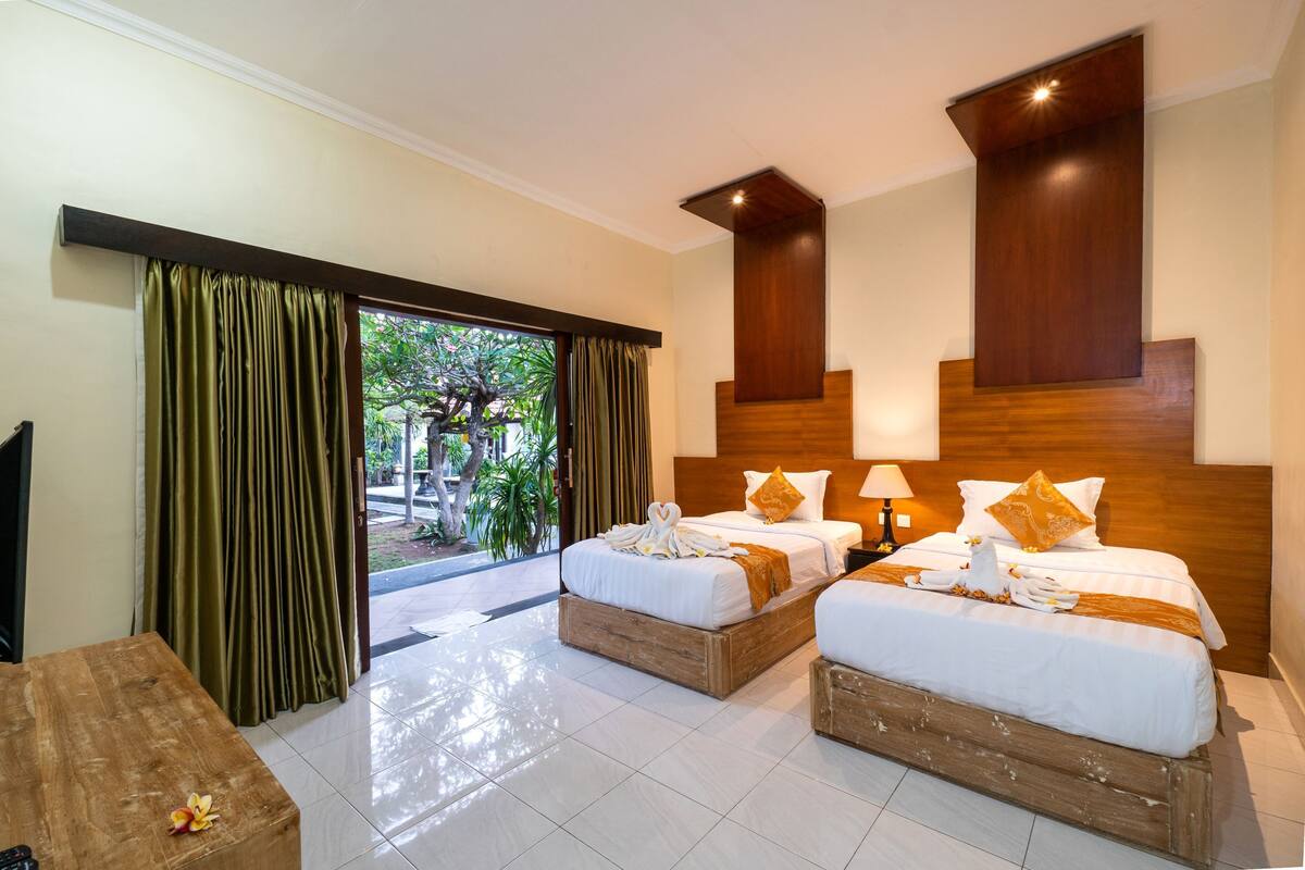 A hotel room with two beds, a wooden headboard, a wooden bench, and a view of greenery outside.