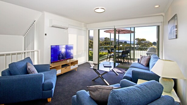 A modern living room with a TV, sofas, and a glass door leading to an outdoor area with a table and chairs. - Anchorage Motel and Villas (Lorne)