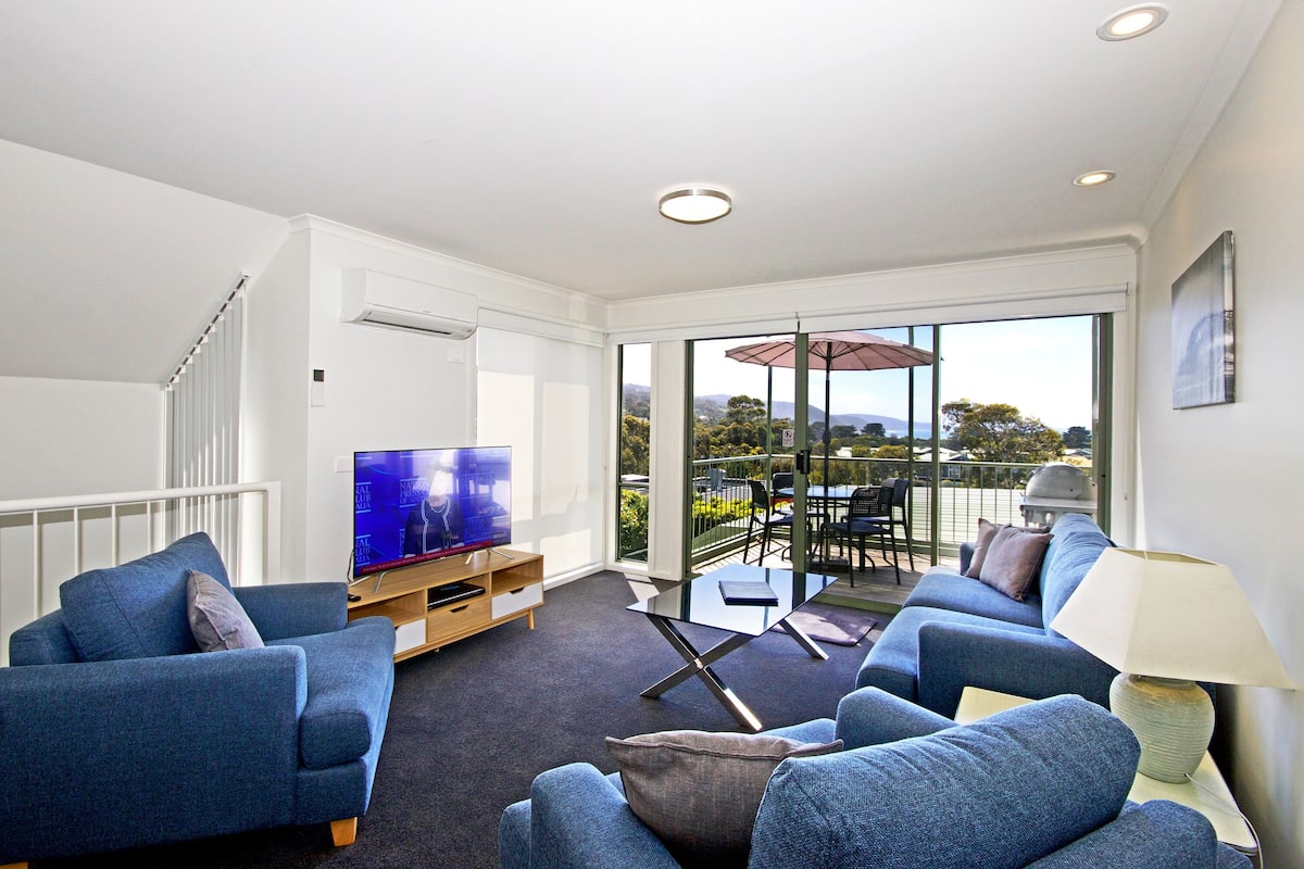 A modern living room with a TV, sofas, and a glass door leading to an outdoor area with a table and chairs.