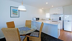 A modern kitchen with white cabinets, a microwave, and a refrigerator. There is a glass dining table with wicker chairs. - Anchorage Motel and Villas (Lorne)