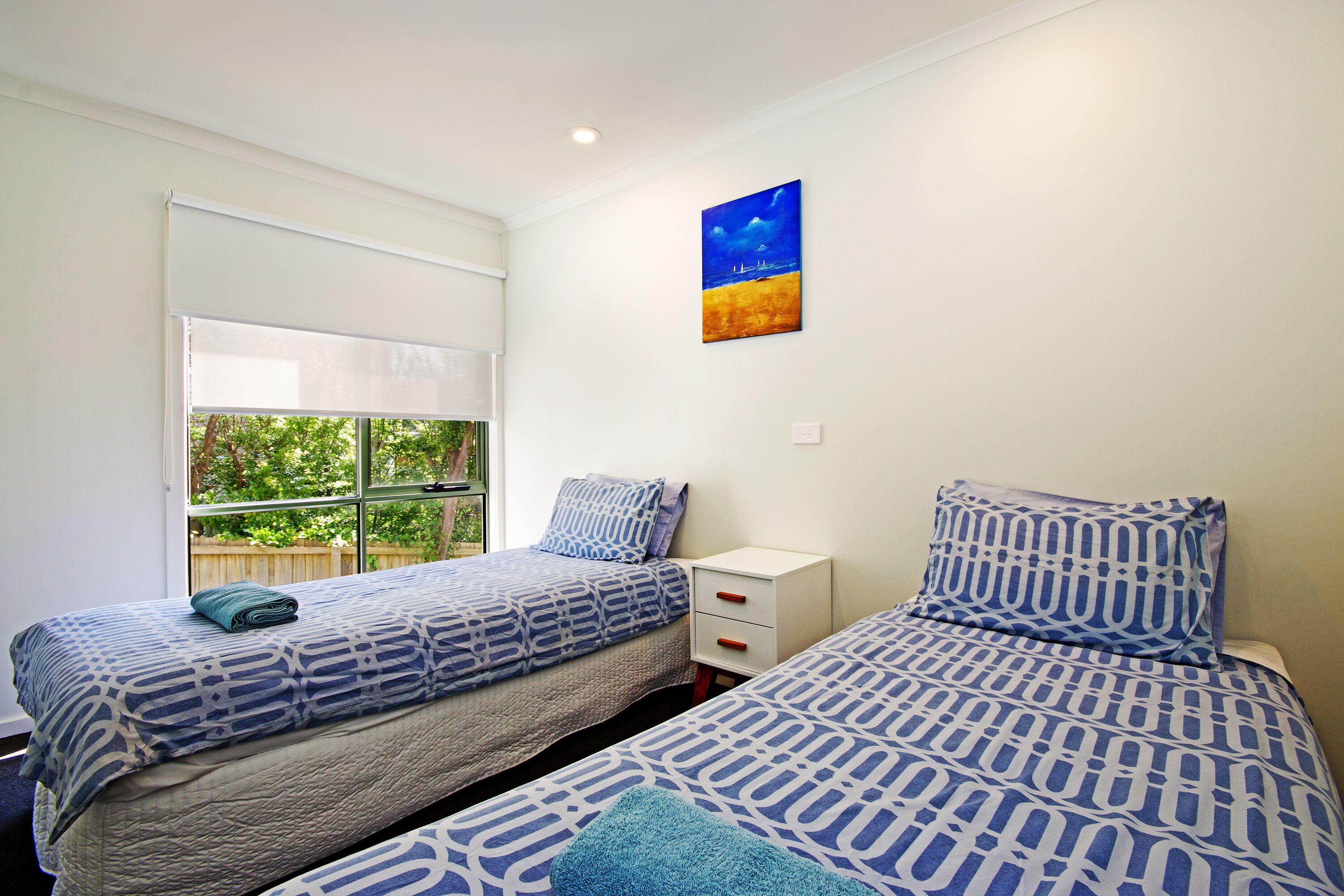 A bedroom with two beds, a window with blinds, a nightstand, and a painting on the wall.