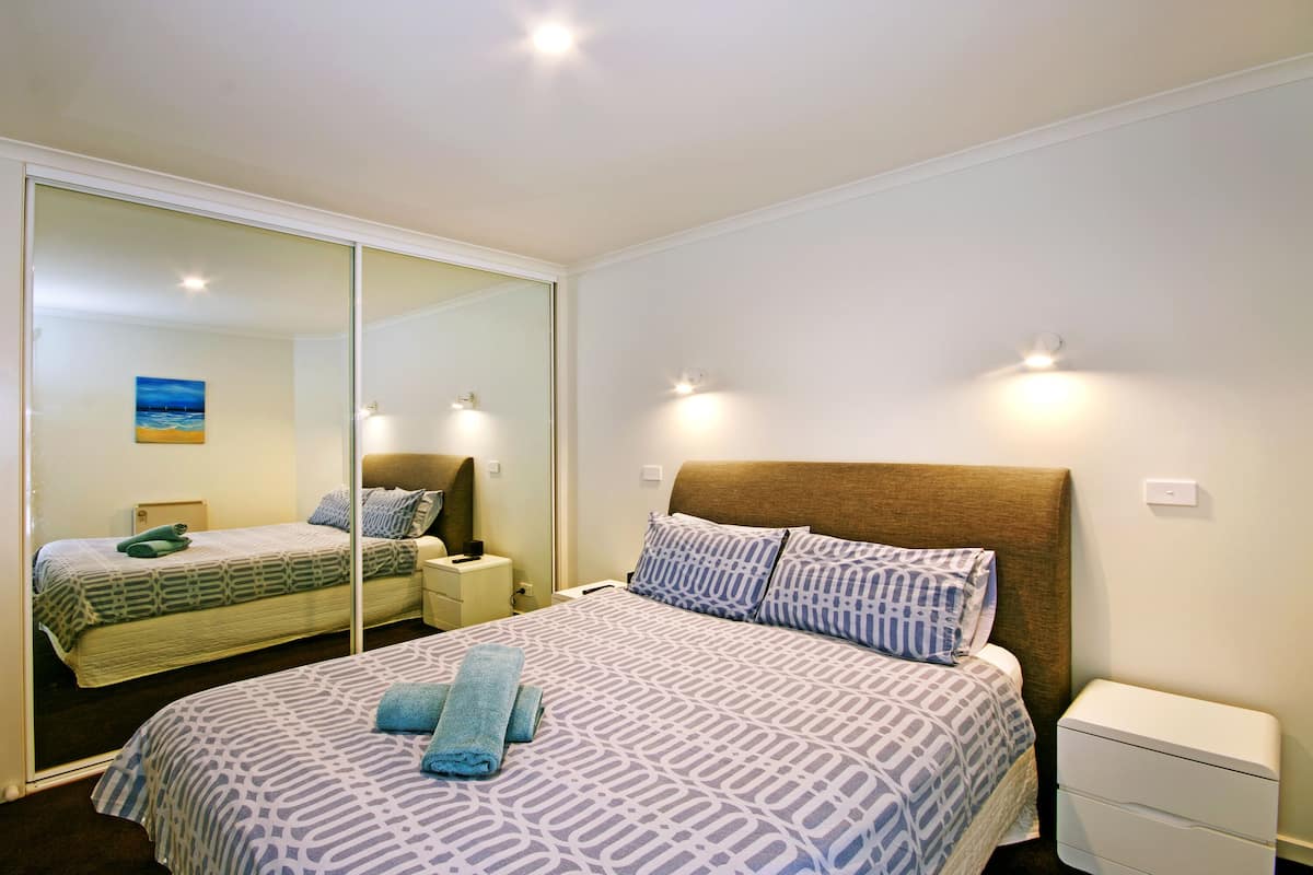 A hotel room with a large bed, a bedside table, a nightstand, a mirror, and a wall-mounted light fixture.