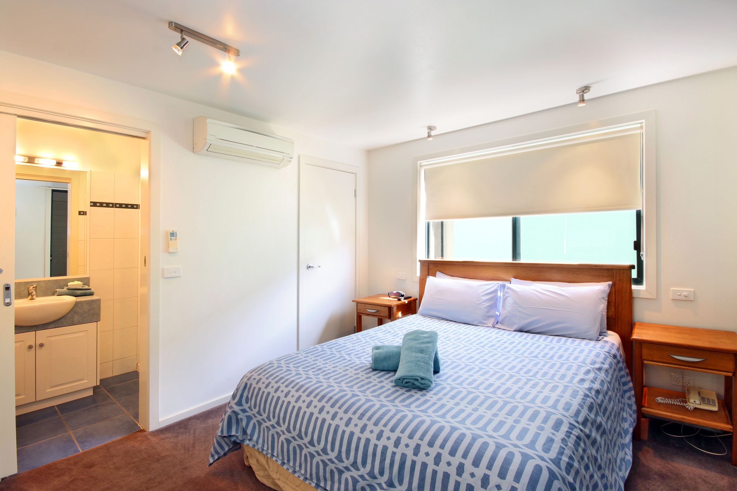Una habitación de hotel con cama, mesita de noche, ventana con persianas, aire acondicionado, baño con lavamanos y espejo, y una puerta.