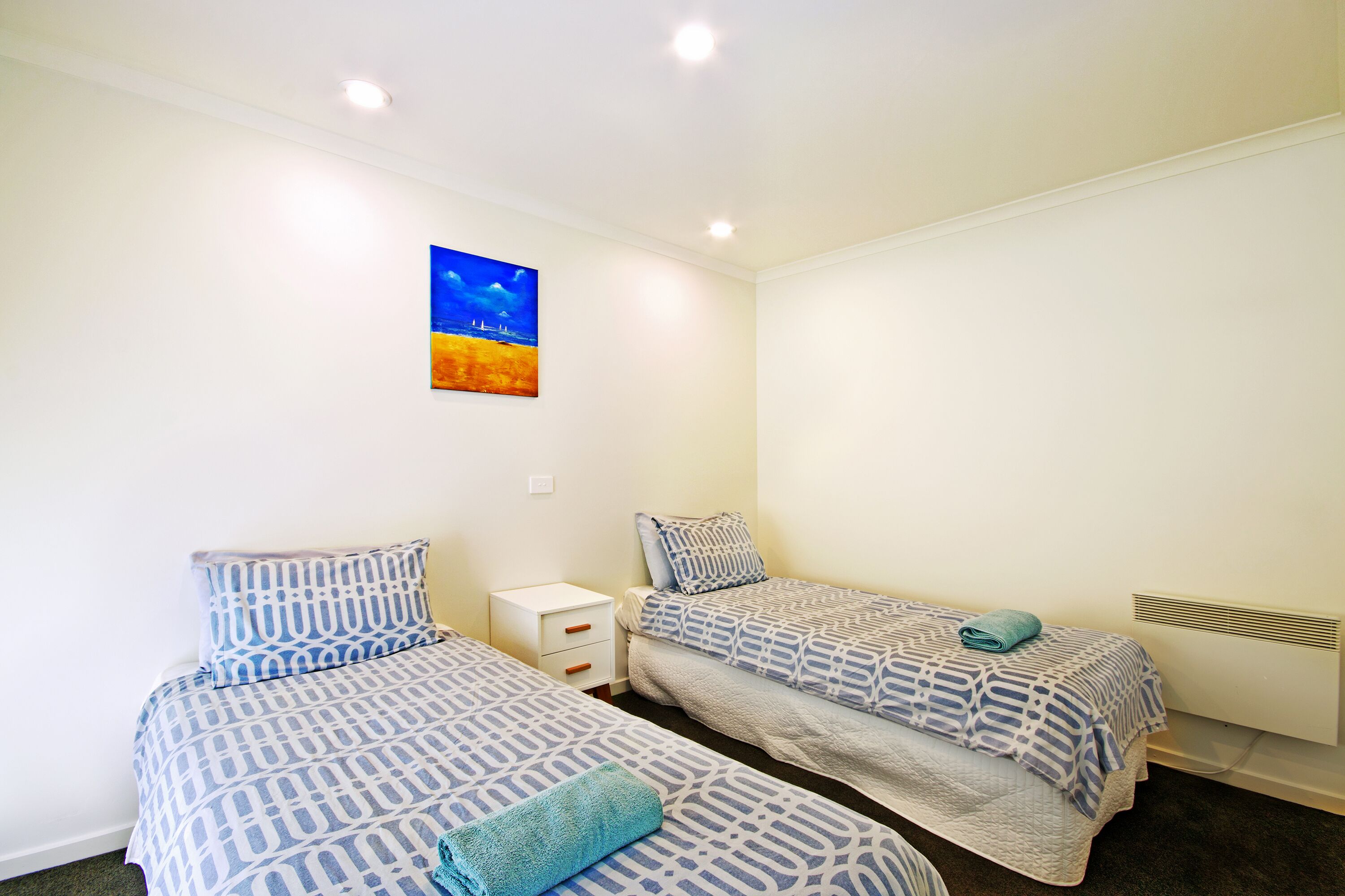 A room with two beds, each with patterned bedding, a small bedside table, and a wall-mounted heater.