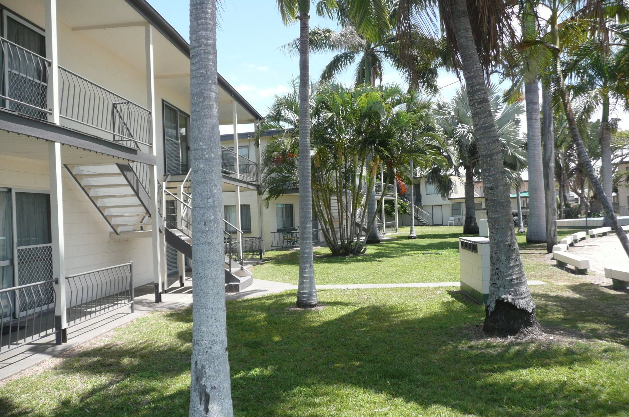 A building with a two-story balcony, a staircase, and a grassy courtyard with palm trees.