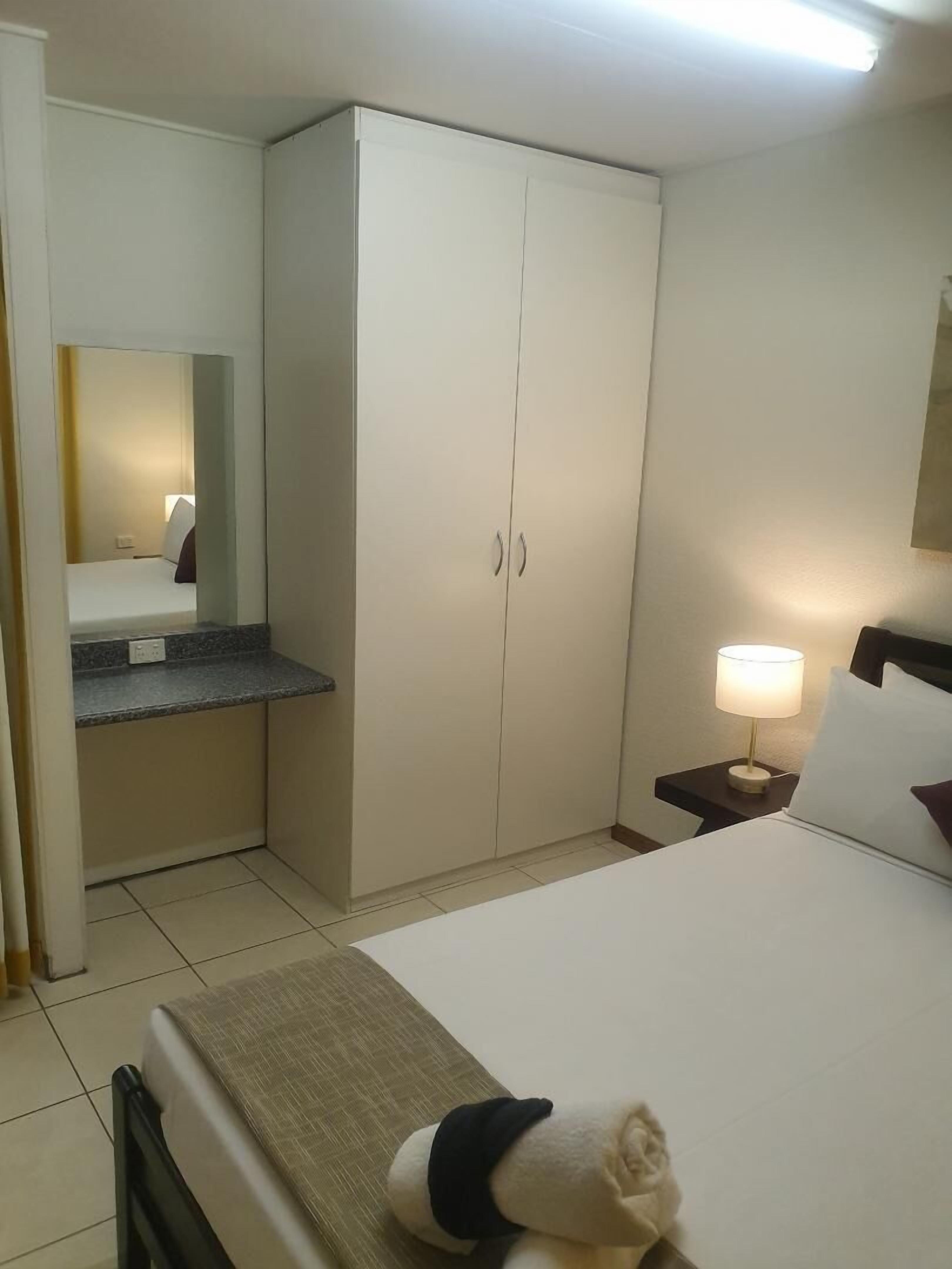 A hotel room with a bed, a nightstand with a lamp, a wardrobe, and a mirror.
