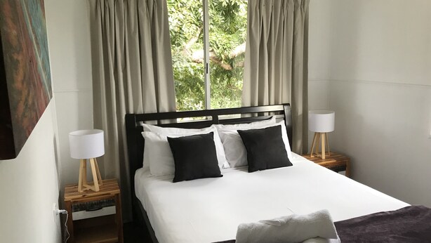 A neatly made bed with white and black bedding, a wooden headboard, and a window with curtains. There are two bedside tables with lamps and a small wooden nightstand. - Ocean Breeze Units (Bowen)