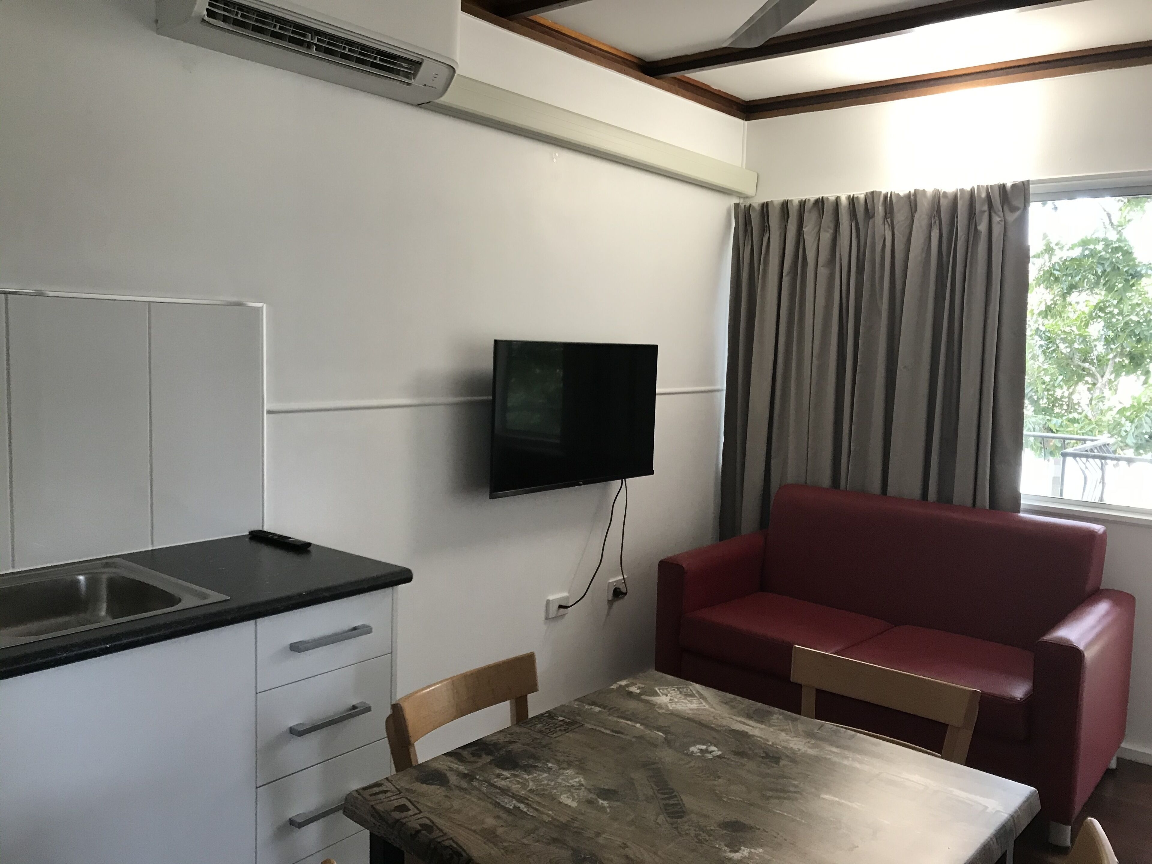 A compact room with a kitchenette, a flat-screen TV mounted on the wall, a dining table with a chair, and a red sofa.
