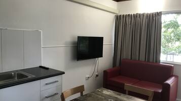 A compact room with a kitchenette, a flat-screen TV mounted on the wall, a dining table with a chair, and a red sofa.