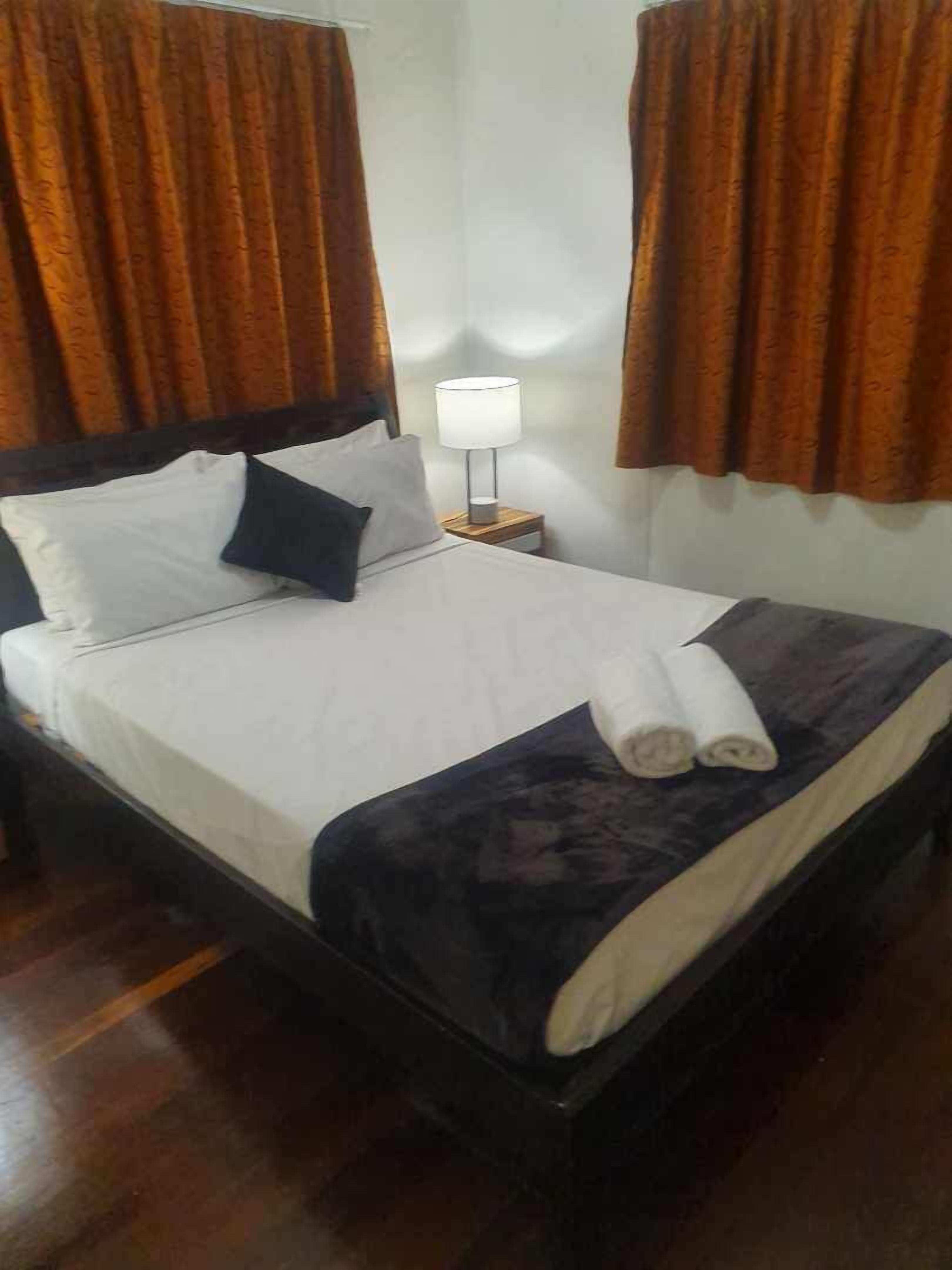 A neatly made bed with white and dark bedding, a small bedside table with a lamp, and brown curtains.