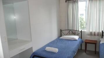 A single bed with a blue mattress, a wooden bedside table, and a window with curtains.