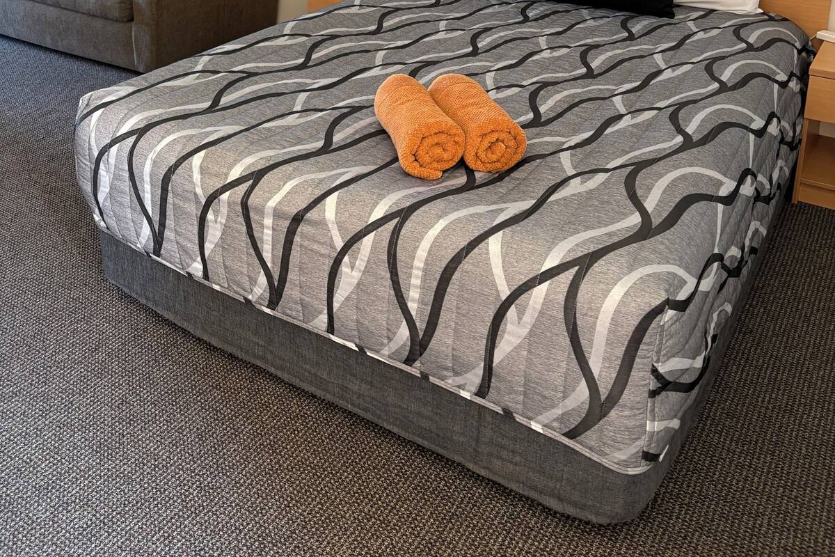 A neatly made bed with a patterned bedspread, a pair of rolled towels, a wooden headboard, a nightstand, a lamp, and a couch with pillows.