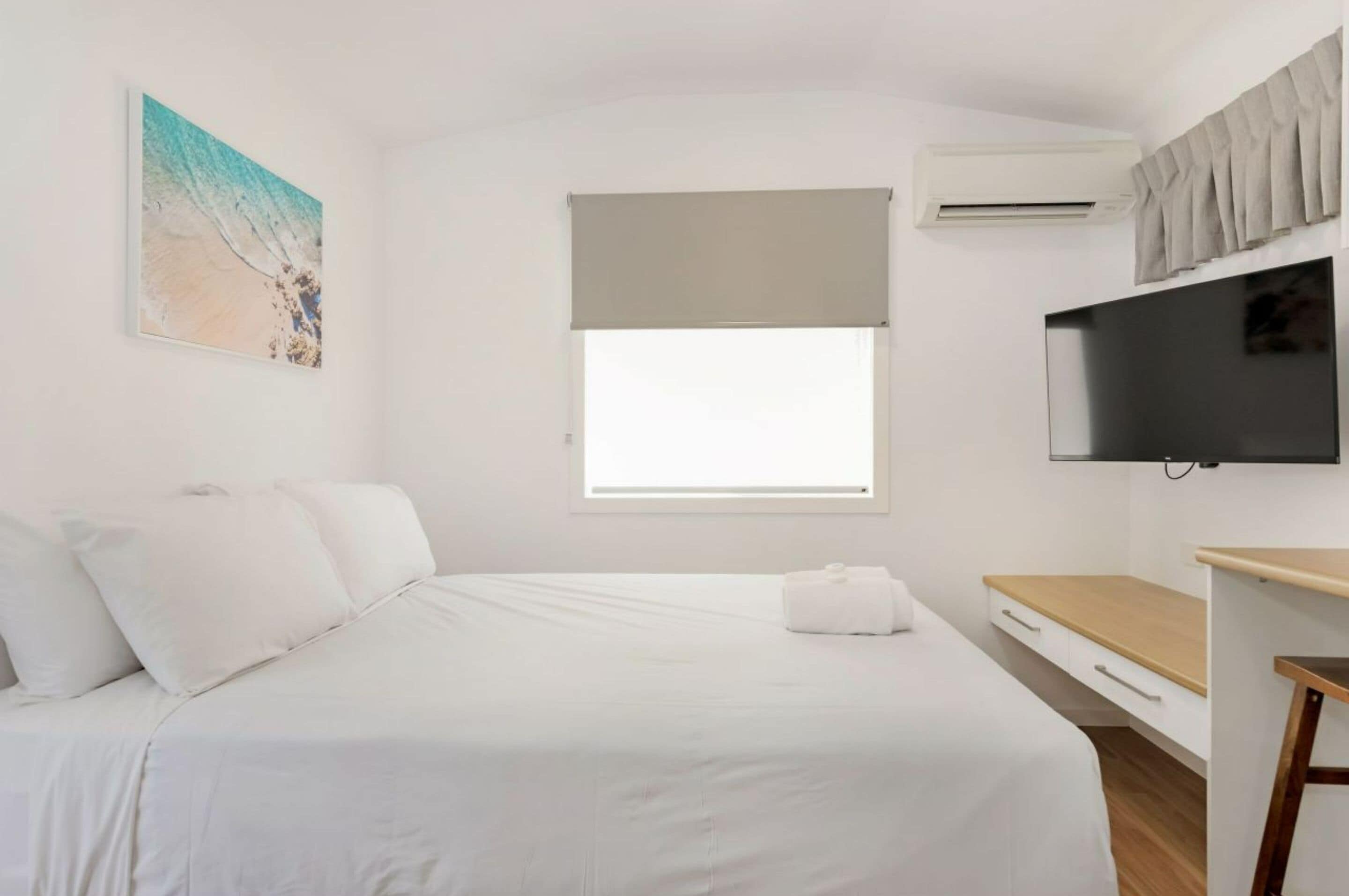 A hotel room with a bed, a flat-screen TV, a window with a beige blind, and an air conditioning unit.
