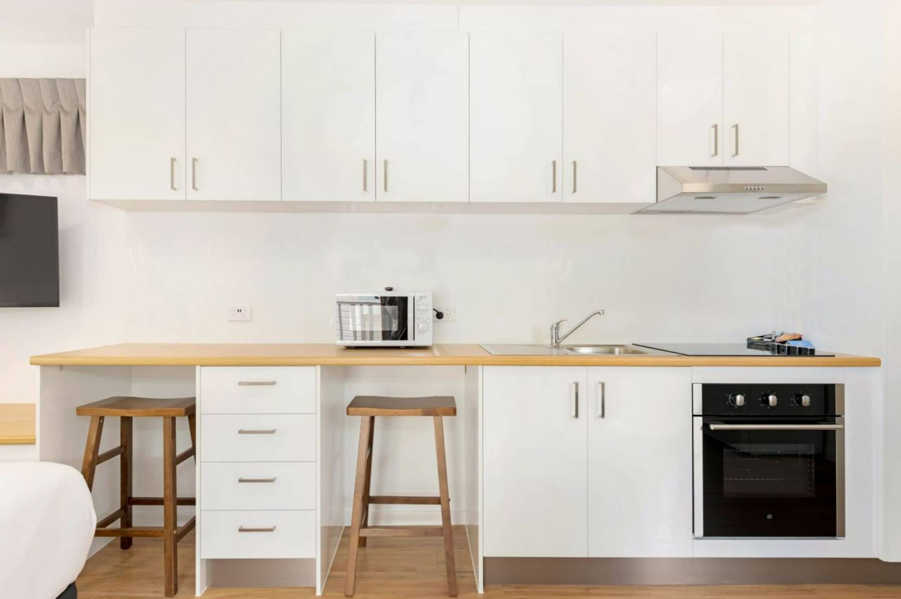A compact kitchen with white cabinets, a microwave, a stove, and two stools.