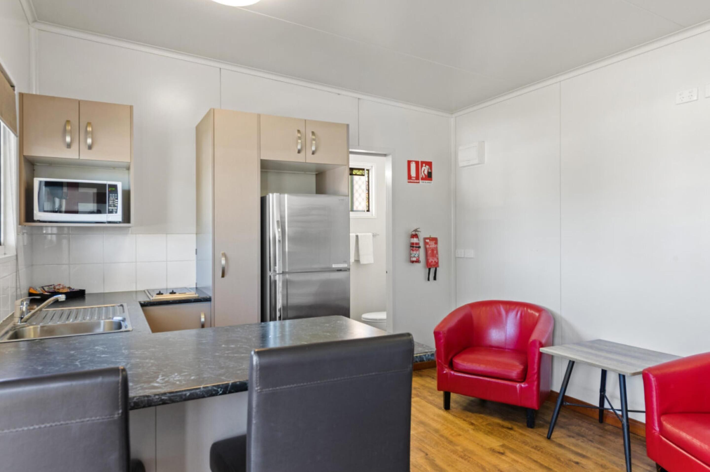 A compact kitchen with a microwave, refrigerator, and sink. There are two red chairs and a small table.