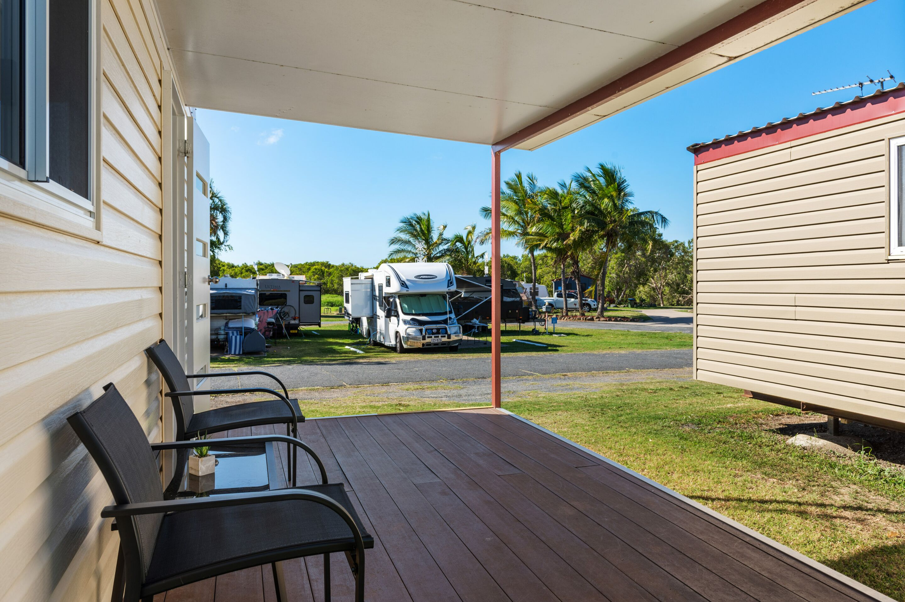 A covered outdoor area with a wooden deck, black chairs, and a view of RVs and a mobile home in the background.