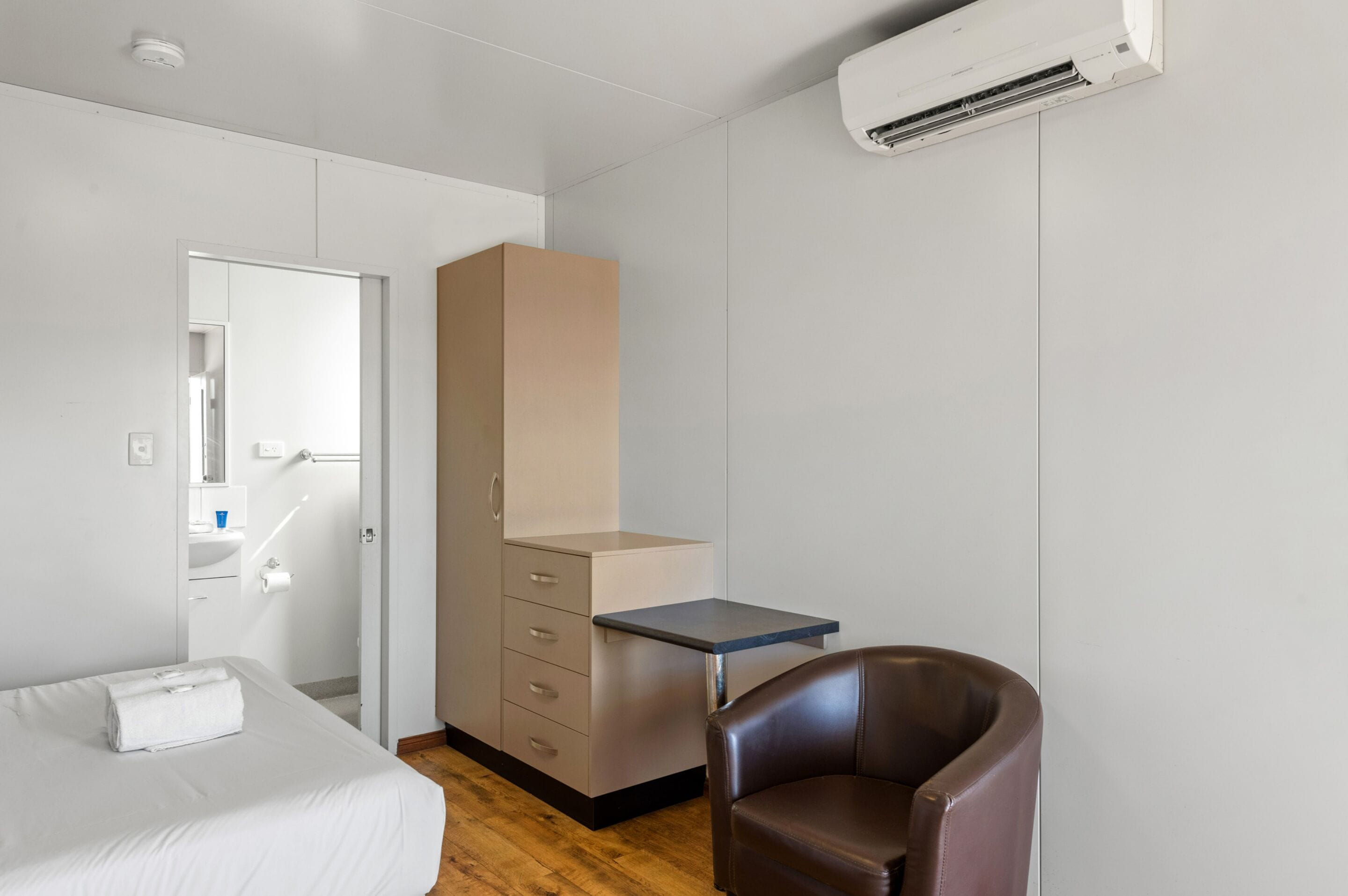 A bedroom with a bed, a desk, a chair, a wardrobe, and an air conditioning unit.