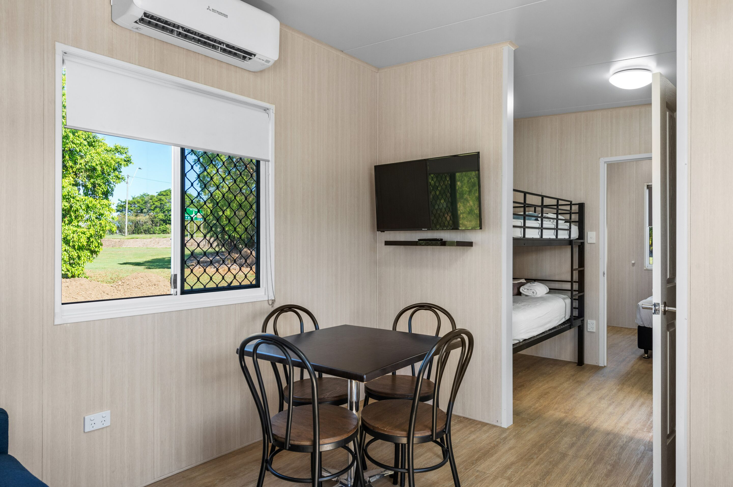 A compact room with a bunk bed, a TV mounted on the wall, a dining table with chairs, and a window with a view of greenery.