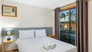 Habitación de hotel con cama, mesita de noche con lámpara, ventana con cortinas y vista a palmeras.