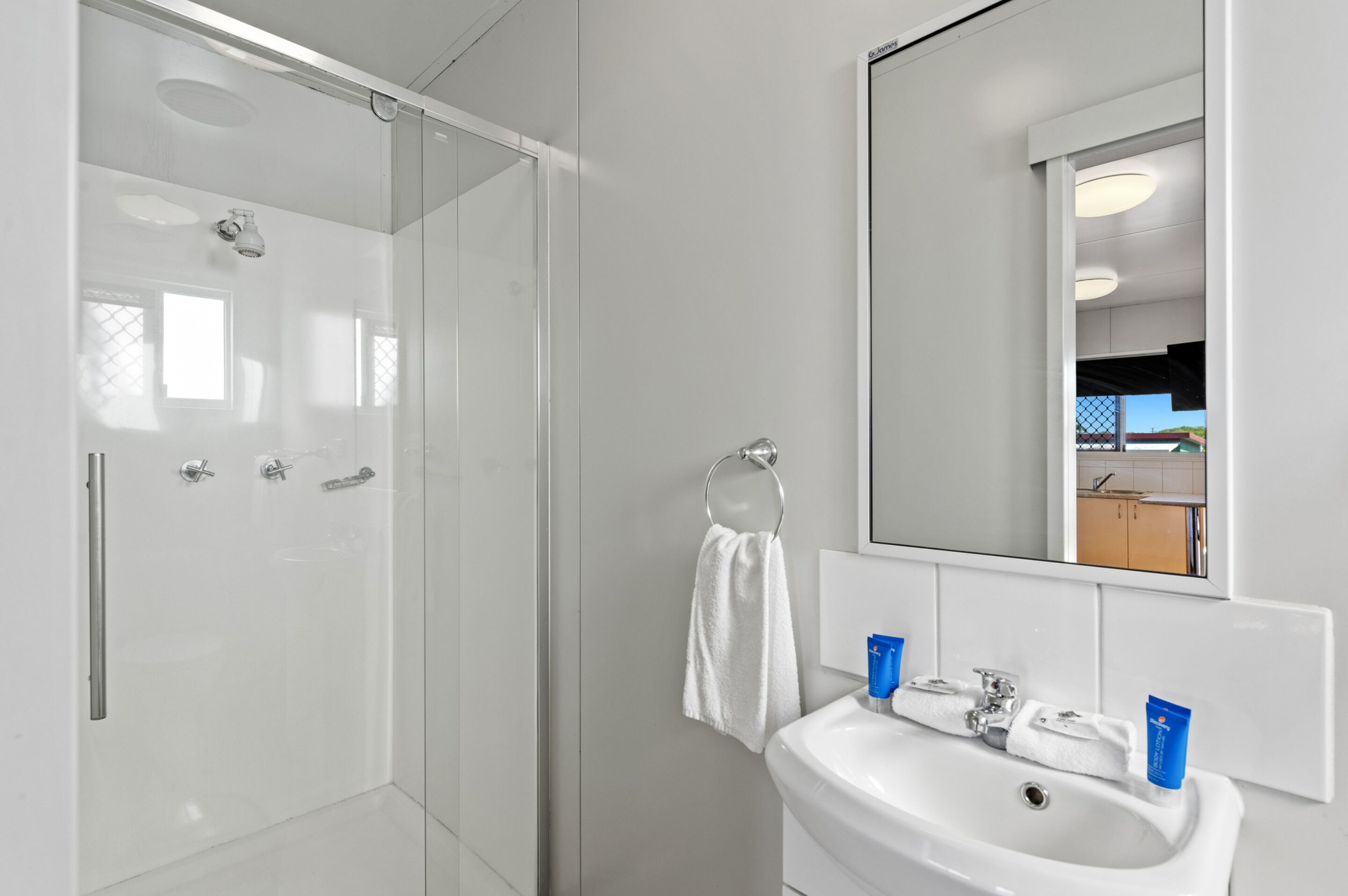 A modern bathroom with a glass shower enclosure, a white sink with two blue soap dispensers, and a large mirror reflecting a kitchen area.