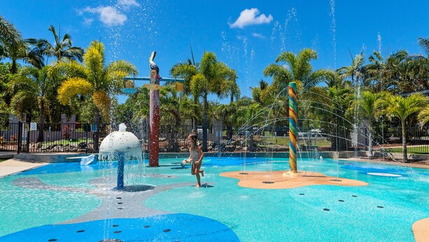 Parque acuático con una fuente central, múltiples chorros de agua y un niño jugando.
