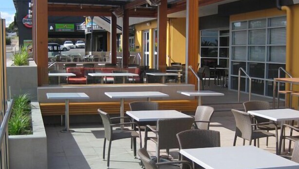 An outdoor patio area with tables and chairs, a covered seating area, and a view of the street in the background. - Christies Beach Hotel (Christies Beach)