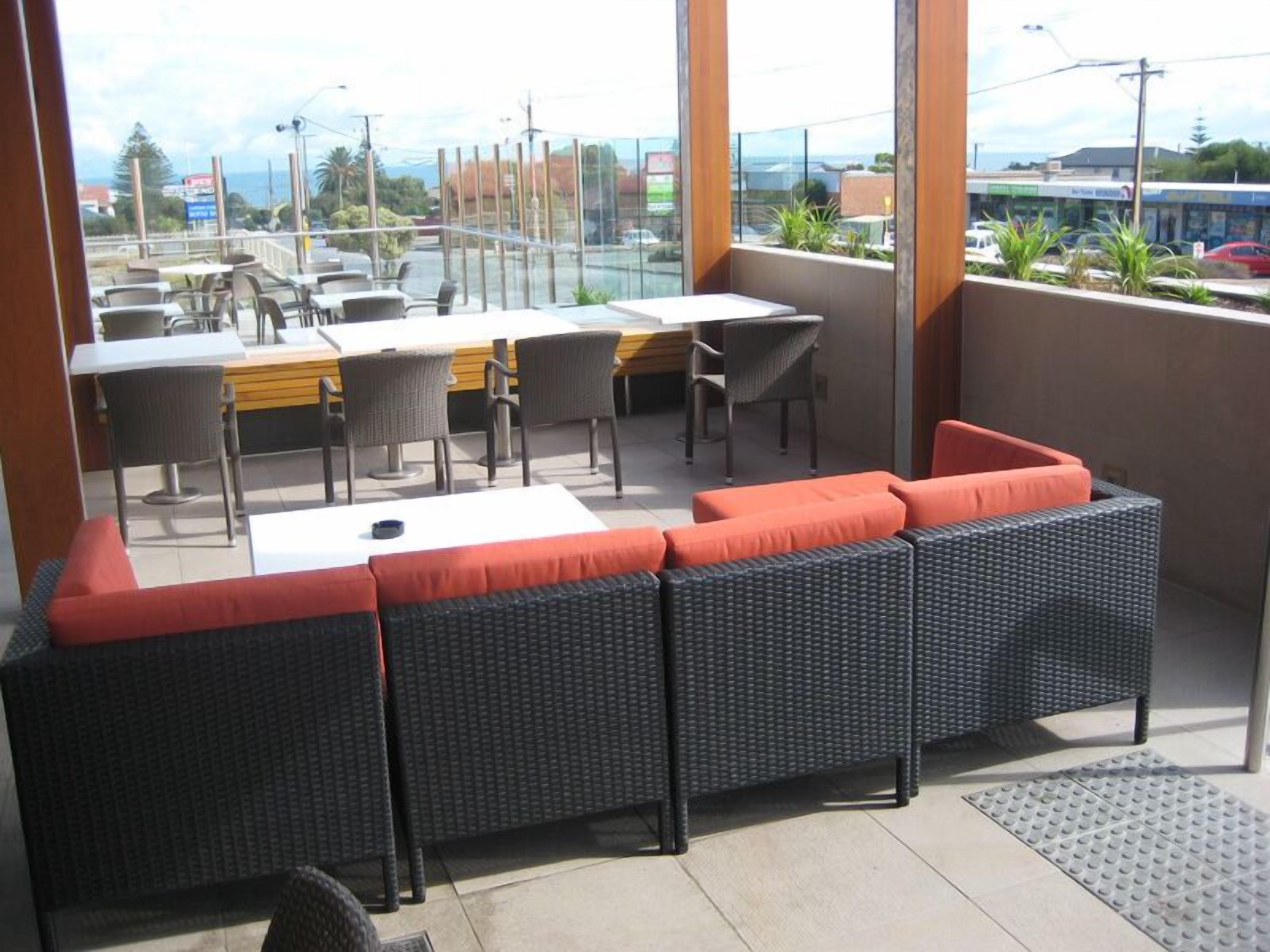 A balcony with wicker furniture, including a sofa and tables, overlooking a street with buildings and a clear sky.