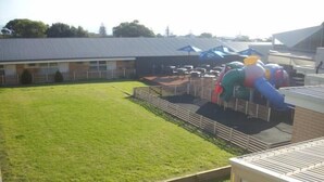 A grassy outdoor area with a fenced play structure, a building with a sloped roof, and a parking lot with cars. - Christies Beach Hotel (Christies Beach)
