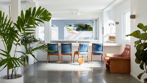 A modern kitchen with a dining area, featuring a large window, potted plants, and a mix of blue and brown chairs. - The Sails Motel Brunswick Heads (Brunswick Heads)