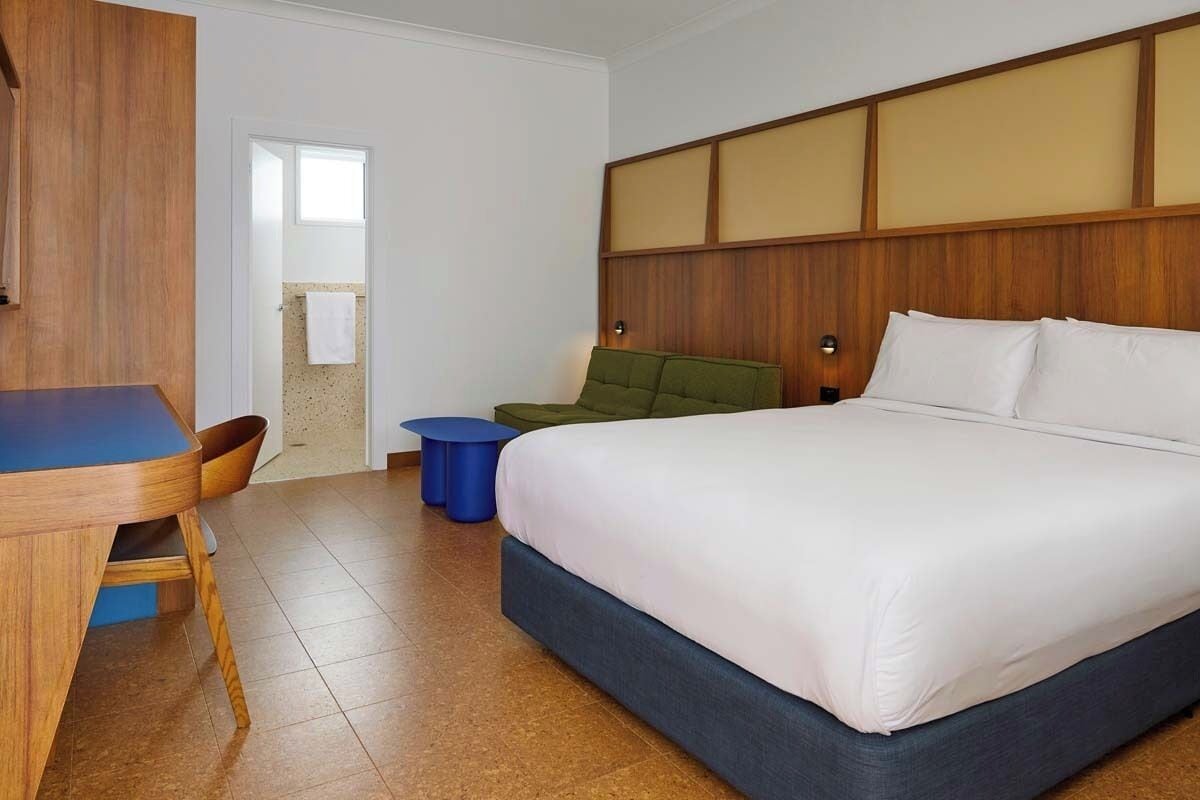A hotel room with a wooden headboard, a bed with white linens, a desk with a chair, and a blue side table.