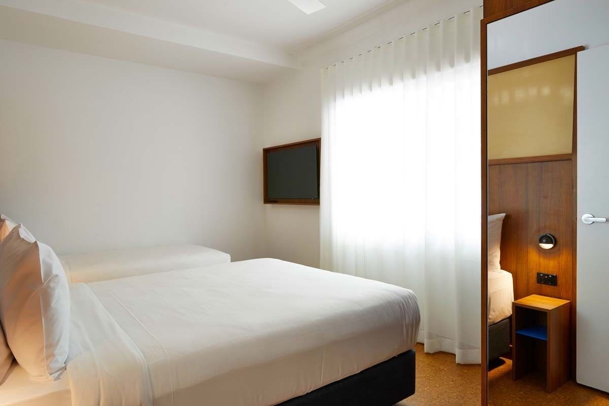 Quarto de hotel com duas camas, uma mesinha de cabeceira, uma pequena escrivaninha, uma cadeira, uma janela com cortinas e uma porta com interruptor de luz.