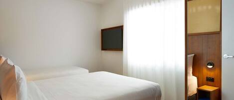 A hotel room with two beds, a bedside table, a small desk, a chair, a window with curtains, and a door with a light switch.