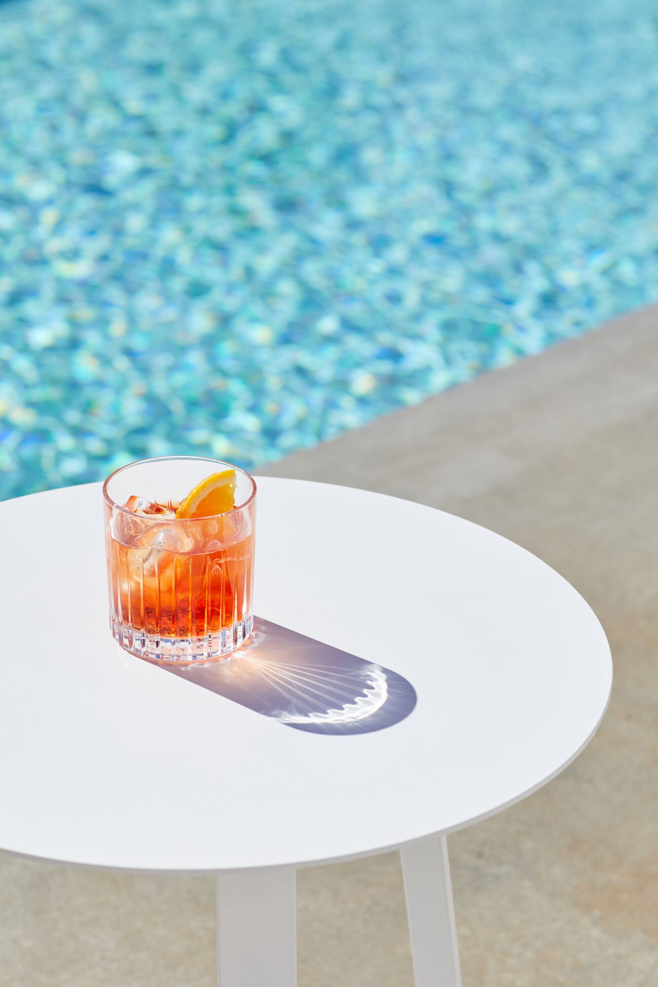 Et drikkeglas med et appelsinstykke på et hvidt bord ved en pool.