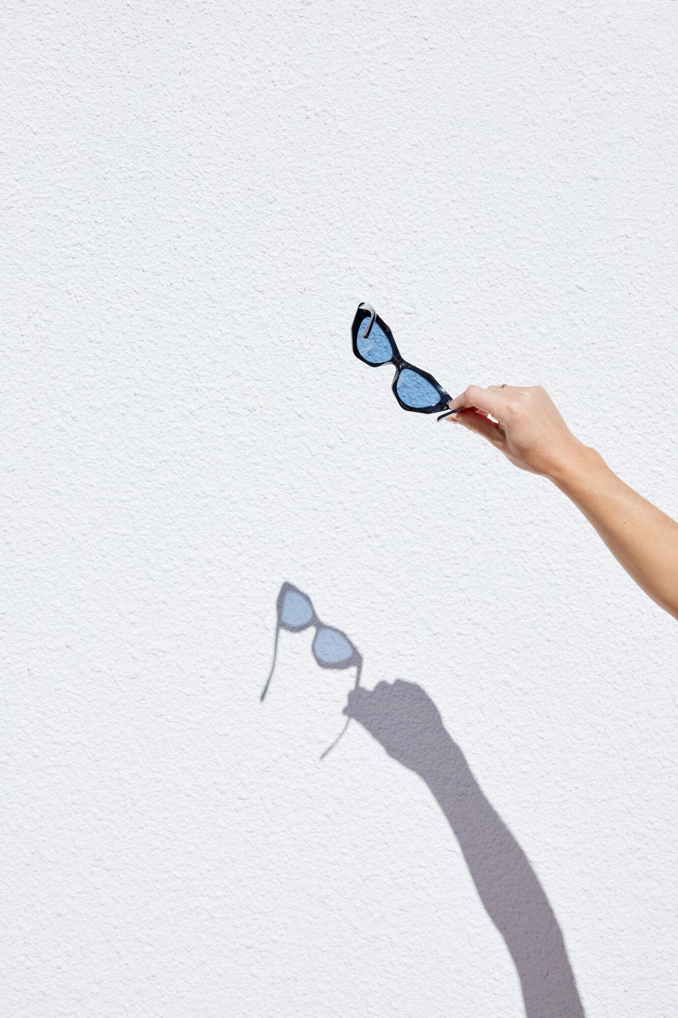 A hand holding sunglasses against a white textured wall.